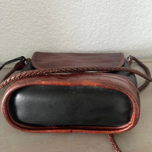 May include: A brown and black leather handbag with a braided strap. The bag has a flap closure and a textured crocodile pattern.