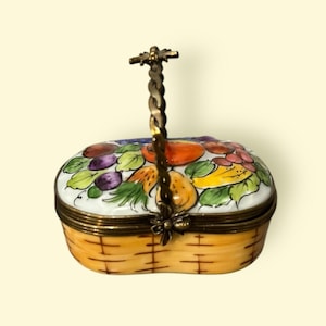 Könnte beinhalten: Eine kleine, ovale Porzellan-Schmuckschatulle in Form eines Korbes mit einem Messinggriff. Der Deckel ist mit einer farbenfrohen Malerei von Früchten verziert, darunter Trauben, Birnen, Bananen und eine große Orange. Der Boden der Schachtel ist so bemalt, dass er wie ein geflochtener Korb aussieht.