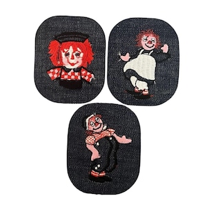 Peut inclure: Trois écussons brodés avec un motif de poupée Raggedy Ann. Chaque écusson est de forme ovale en denim bleu foncé. La poupée a des cheveux en fil rouge, un visage blanc et un chapeau noir. La poupée porte un haut à carreaux rouges et blancs.