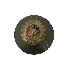 May include: A round, dark gray marble bottle stopper with a gold-colored, ornate, circular design. The gold design features a central floral element surrounded by a detailed, repeating pattern. The stopper is designed to fit a bottle.