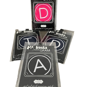 Puede incluir: Cuatro parches de monograma para despegar y pegar en diferentes colores y letras. Los parches están empaquetados en bolsas de plástico transparentes con etiquetas en blanco y negro que dicen "Insta mono GRAM" y "mudpie". Las letras en los parches son D, T, A y D.