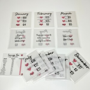 May include: A set of 12 clear plastic envelopes with pink hearts and handwritten text. Each envelope has a month of the year written on it, along with a dollar amount. The envelopes are designed for a savings challenge.