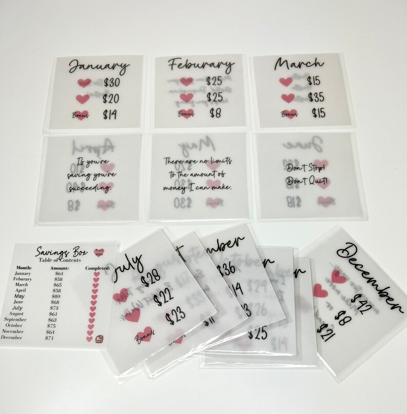 May include: A set of 12 clear plastic envelopes with pink hearts and handwritten text. Each envelope has a month of the year written on it, along with a dollar amount. The envelopes are designed for a savings challenge.