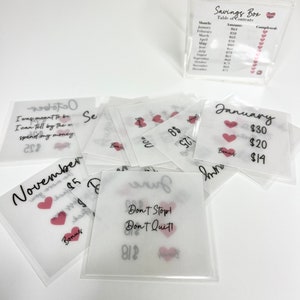 May include: A set of 12 clear envelopes with pink hearts and handwritten text. Each envelope has a month of the year written on it, along with a dollar amount. The envelopes are labeled "Bonus" and are part of a savings box system.