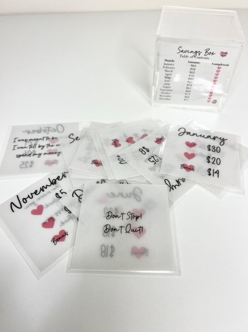 May include: A set of 12 clear envelopes with pink hearts and handwritten text. Each envelope has a month of the year written on it, along with a dollar amount. The envelopes are labeled "Bonus" and are part of a savings box system.