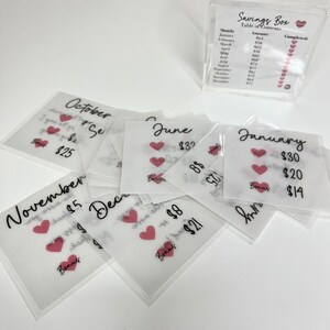 May include: A set of 12 clear envelopes with a pink heart and handwritten text. Each envelope has a month of the year and a dollar amount. The envelopes are stacked on top of each other. The text on the envelopes reads: January $30, $20, $14, June $32, $8, $21, October $25, November $15, December $8, $21.