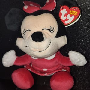 May include: A plush Minnie Mouse Beanie Baby. The doll has a red dress with white polka dots, a red bow with white polka dots, and black ears. The tag reads "Beanie Babies".