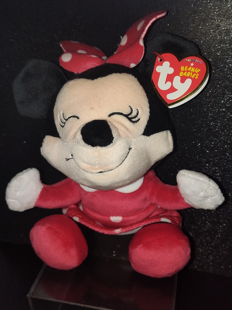 May include: A plush Minnie Mouse Beanie Baby. The doll has a red dress with white polka dots, a red bow with white polka dots, and black ears. The tag reads "Beanie Babies".