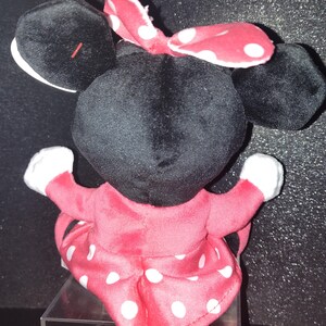 May include: A plush Minnie Mouse doll, viewed from the back. The doll has a black head and ears, a red and white polka dot bow, and a red dress with white polka dots. The doll's arms are outstretched, with white gloves.