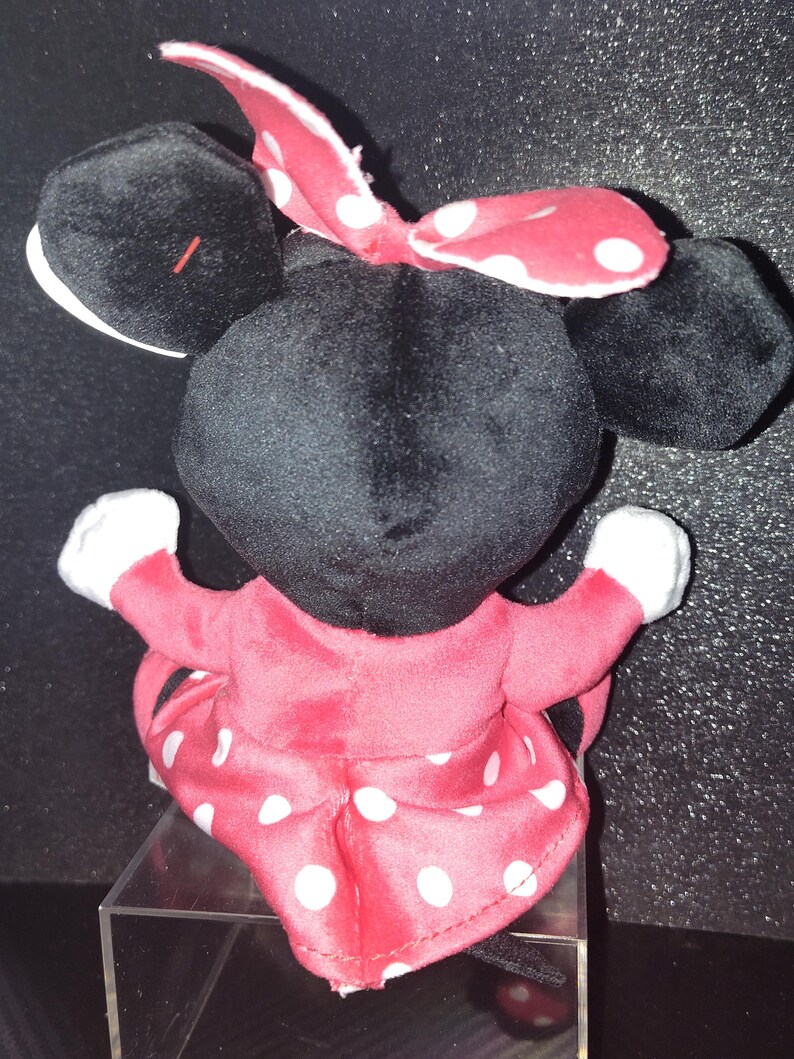 May include: A plush Minnie Mouse doll, viewed from the back. The doll has a black head and ears, a red and white polka dot bow, and a red dress with white polka dots. The doll's arms are outstretched, with white gloves.