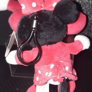 May include: A plush Minnie Mouse keychain. The toy features a red dress with white polka dots, a matching bow, and black ears. The keychain has a black clasp for attaching to keys or bags. The plush toy is soft and cuddly.