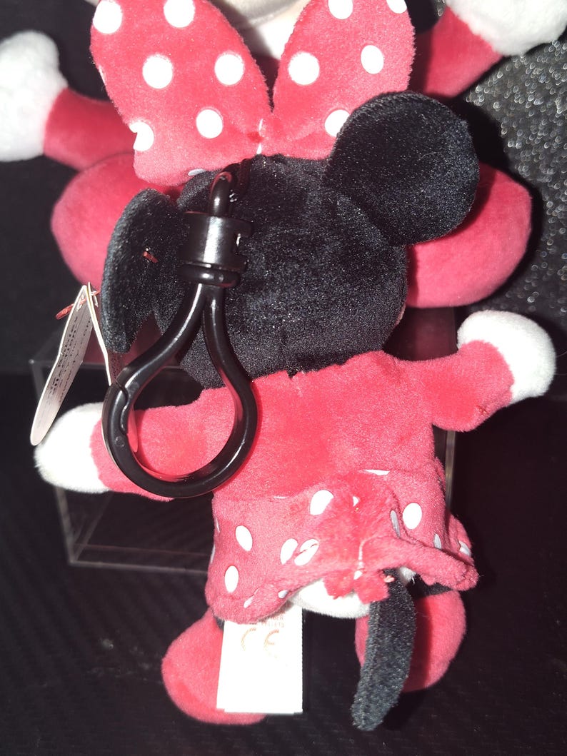 May include: A plush Minnie Mouse keychain. The toy features a red dress with white polka dots, a matching bow, and black ears. The keychain has a black clasp for attaching to keys or bags. The plush toy is soft and cuddly.