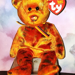 May include: A plush teddy bear with a mottled orange and black pattern, featuring a red heart-shaped Ty Beanie Babies tag, a small MasterCard logo on its chest, and a ribbon around its neck with black, red, and yellow stripes.