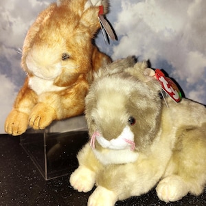 May include: Two plush rabbit toys. One is light brown with a cream-colored face and paws. The other is a mix of tan and brown. Both have red tags attached to their ears.