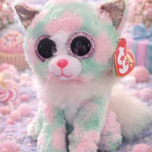 May include: A plush cat toy in pastel colors of pink and mint green, with glittery accents on the ears and around the eyes. The cat has large, reflective eyes and a pink nose. A red heart-shaped tag is attached to the toy.