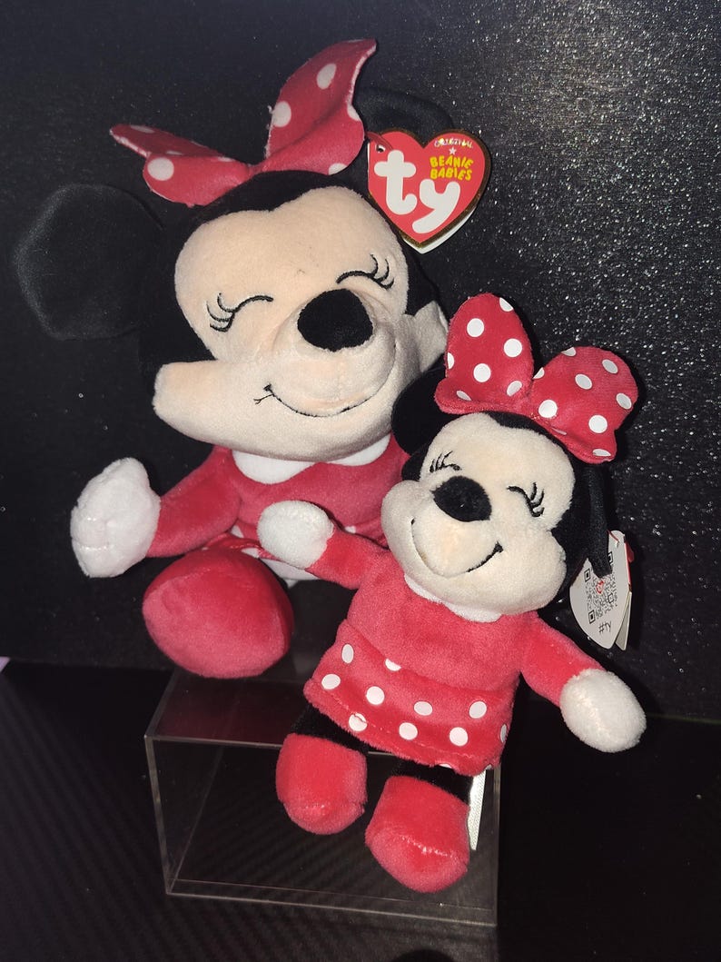 May include: Two Minnie Mouse plush toys. The larger one has a red dress and a red bow with white polka dots. The smaller one has a red dress with white polka dots and a matching bow. Both have black ears and closed eyes.
