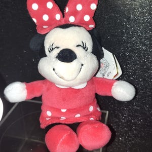 May include: A plush Minnie Mouse doll in a red dress with white polka dots and a matching bow. The doll has a black nose, black ears, and a smiling face. The doll's arms and feet are red.