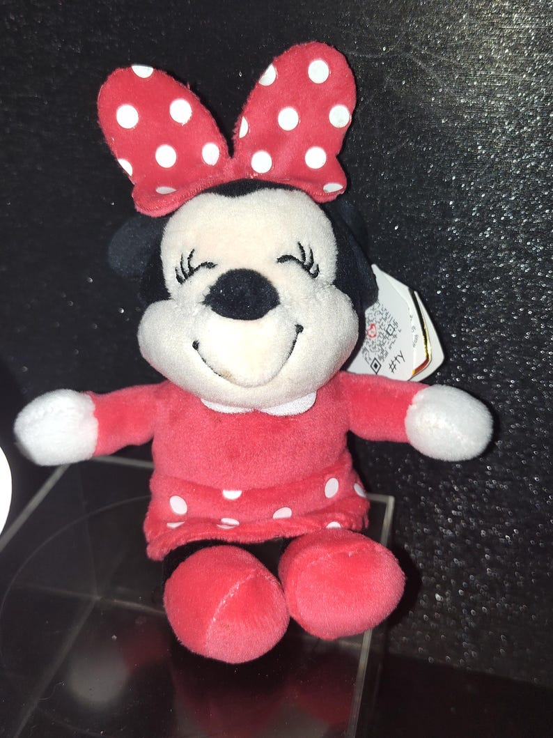 May include: A plush Minnie Mouse doll in a red dress with white polka dots and a matching bow. The doll has a black nose, black ears, and a smiling face. The doll's arms and feet are red.