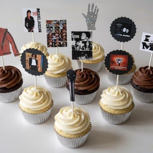 May include: A collection of cupcakes in silver foil liners topped with white or chocolate frosting and decorative paper toppers featuring Michael Jackson imagery, including a red jacket, a glittery glove, a microphone, and text like I Love MJ.
