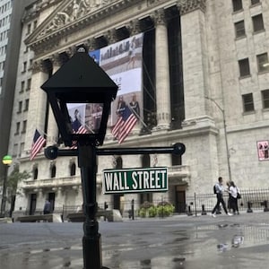 Peut inclure: Un lampadaire noir avec une lanterne noire sur le dessus, sur lequel est fixé un panneau de rue noir avec une inscription blanche "WALL STREET". Le lampadaire est situé devant un grand bâtiment à façade en pierre.