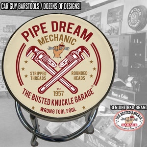 May include: A round bar stool with a cream-colored seat featuring a red and white logo that reads "Pipe Dream Mechanic" with a pair of crossed wrenches and the text "The Busted Knuckle Garage Est. 1957 Wrong Tool Fool".