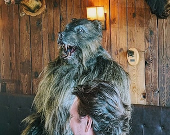May include: A large, furry, gray wolf-like creature sits in a wooden bar setting. A man with dark hair sits next to the creature. The creature has a menacing expression with sharp teeth.
