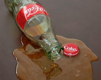 May include: A clear glass Coca-Cola bottle lies on its side, with a red and white label. A spilled brown liquid surrounds the bottle and a red bottle cap with the word "Coke" rests nearby. The scene suggests a beverage mishap.