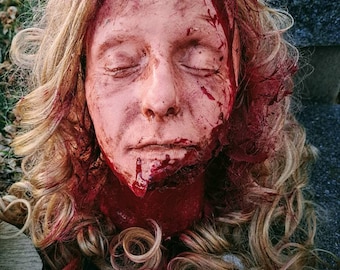 May include: A realistic-looking, bloody, severed head with long, curly, blonde hair. The head is lying on a dark surface.