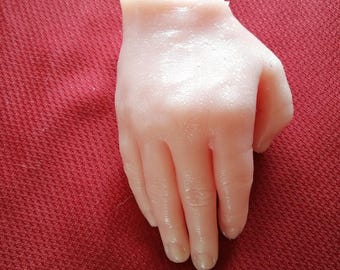 May include: A lifelike, pale pink prosthetic hand with visible knuckles and fingernails. The hand is cut off at the wrist and rests on a textured, dark red surface. The fingers are slightly curved.