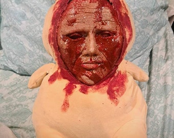 May include: A plush toy shaped like a small animal with a human face. The face is covered in red paint, resembling blood. The toy is lying on a bedspread with a floral pattern.