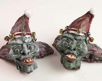 May include: Two green and gray Christmas elf ornaments with red and white Santa hats and gold jingle bells. The elves have a mischievous expression on their faces.