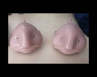 May include: Two pale pink, rubbery, fish-shaped toys with large, round eyes and a downturned mouth. The toys are sitting on a white surface.