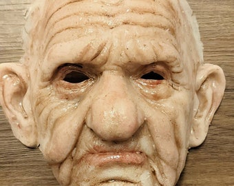 May include: A realistic-looking, detailed, pale-colored, old man's face mask. The mask has wrinkles and a textured surface.
