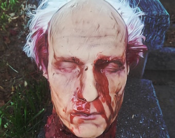 May include: A realistic, detailed, and gruesome prop of a severed head with white hair and a bloody neck. The head is lying on a dark, textured surface.