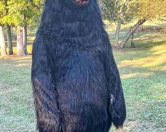 May include: A full-body black bear costume with a detailed face and fur texture. The bear is standing outdoors in a grassy area with trees in the background. The costume has visible teeth and orange eyes.