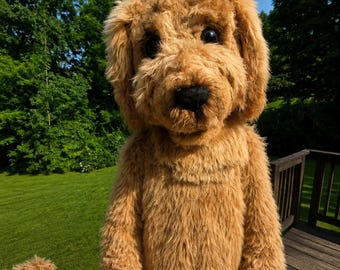 May include: A full-body mascot costume designed to resemble a golden-colored dog with shaggy, textured faux fur. The costume features a large, rounded head with floppy ears, dark round eyes, and a black nose, with a matching furry body and arms.
