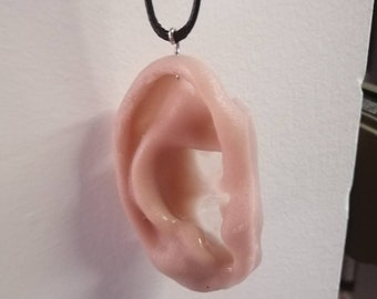 May include: A light pink ear-shaped pendant hangs from a black cord necklace. The pendant is made of a smooth, slightly textured material.