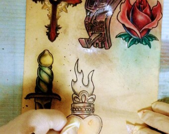 May include: A sheet of paper with several tattoo designs drawn on it. The designs include a cross, a rose, a dagger, a heart with flames, and a geometric design. The designs are drawn in black ink and colored in with red, green, yellow, and blue.