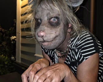 May include: A person wearing a gray rat mask with large ears and a pink nose. The person is wearing a black and white striped shirt and a silver chain necklace. The person is outside at night.