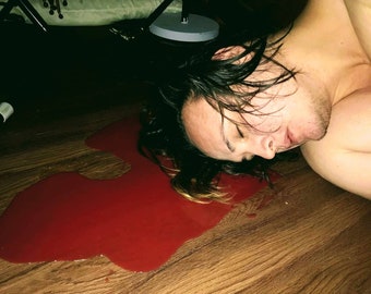May include: A person lies on a wooden floor with a large pool of red liquid around them.