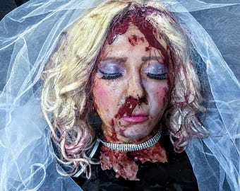 May include: A woman's head with blonde hair, wearing a white veil, covered in blood. The woman has purple eyeshadow and pink lipstick. She is wearing a silver necklace.