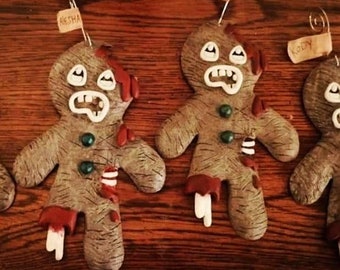 May include: Four gingerbread men ornaments with personalized names, "Aesha", "Bill", "Kody", and an unreadable name. The gingerbread men are decorated with brown, white, and red paint to look like zombies.