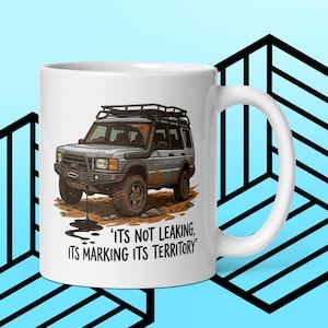 May include: White ceramic mug with a cartoon illustration of a silver SUV with a roof rack and off-road tyres. The text on the mug reads, "ITS NOT LEAKING, ITS MARKING ITS TERRITORY."