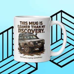 May include: White ceramic mug with a handle. The mug features a graphic of a muddy off-road vehicle and the text: "THIS MUG IS CLEANER THAN MY DISCOVERY. (And that's saying something.)" The background is light blue with black geometric shapes.