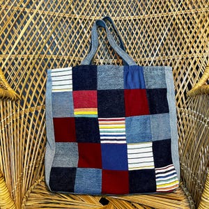 Op de afbeelding: Een vierkante draagtas gemaakt van een patchwork van verschillende stoffen vierkanten, waaronder donkerblauwe denim, effen rood, effen blauw en witte stof met horizontale veelkleurige strepen, voorzien van twee bijpassende denim schouderbanden.