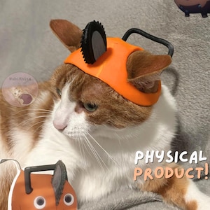 May include: A cat wearing an orange and black chainsaw-themed hat, with a cartoon illustration of a chainsaw dog. The image includes the text "string included!" and "PHYSICAL PRODUCT!"