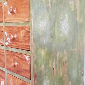 May include: A wooden dresser with multiple drawers painted in a distressed green and white paint. The drawers have a wood grain finish and white floral designs.