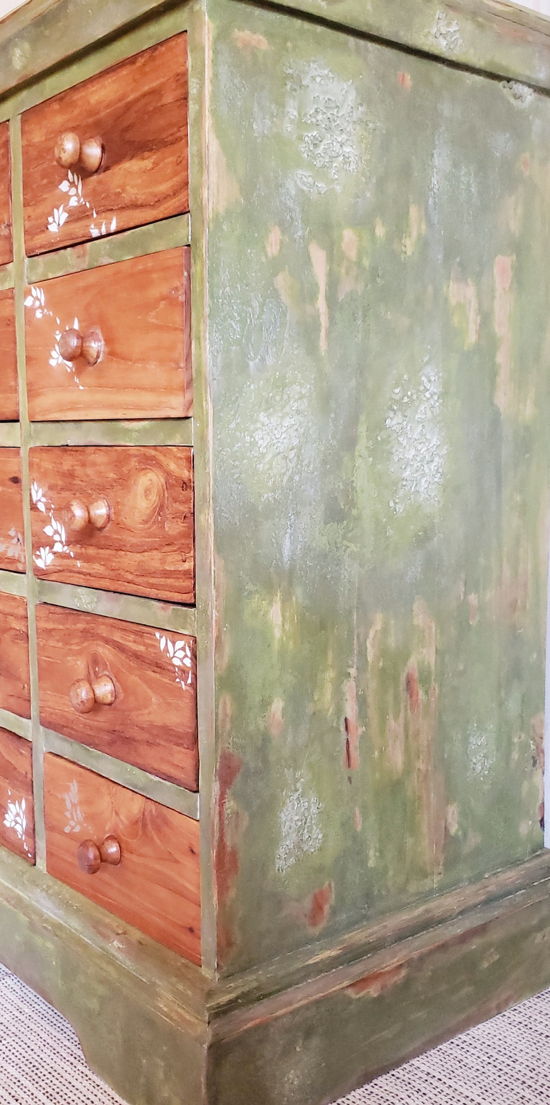 May include: A wooden dresser with multiple drawers painted in a distressed green and white paint. The drawers have a wood grain finish and white floral designs.