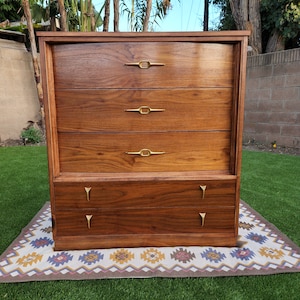 May include: A vintage wooden dresser with five drawers and brass hardware. The dresser has a rich brown wood finish and is sitting on a patterned rug.