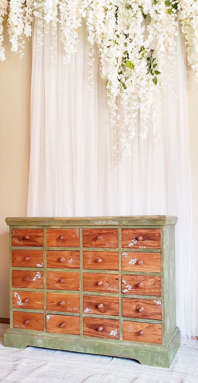 May include: A green painted wooden dresser with 20 drawers. The drawers are made of wood with a natural finish and have white floral designs painted on them. The dresser is sitting on a white rug in front of a white sheer curtain.